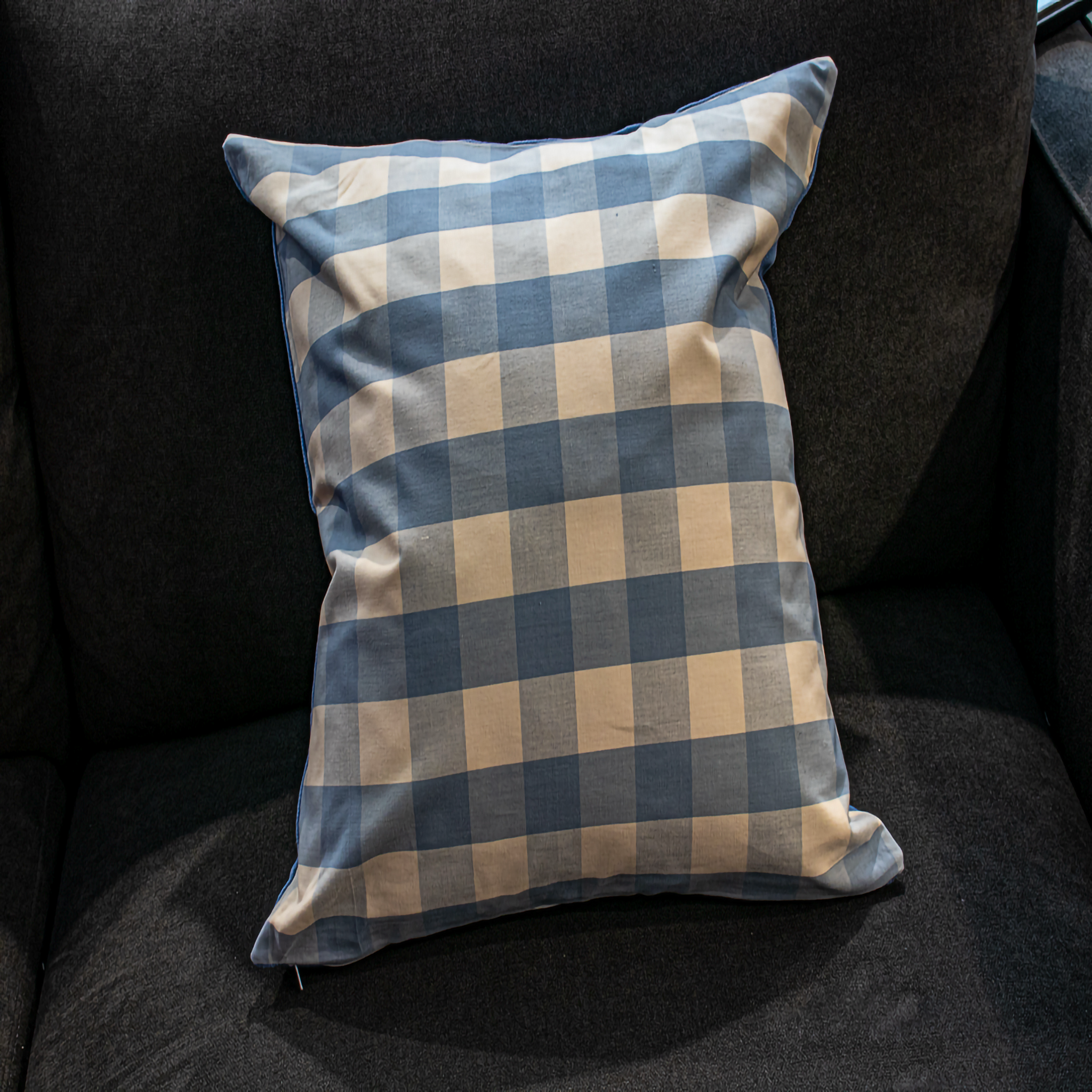 A close-up of the Luxe & Humble Gingham Blue and Beige Pillow featuring a soft cotton texture and a classic checkered gingham pattern on a dark sofa.