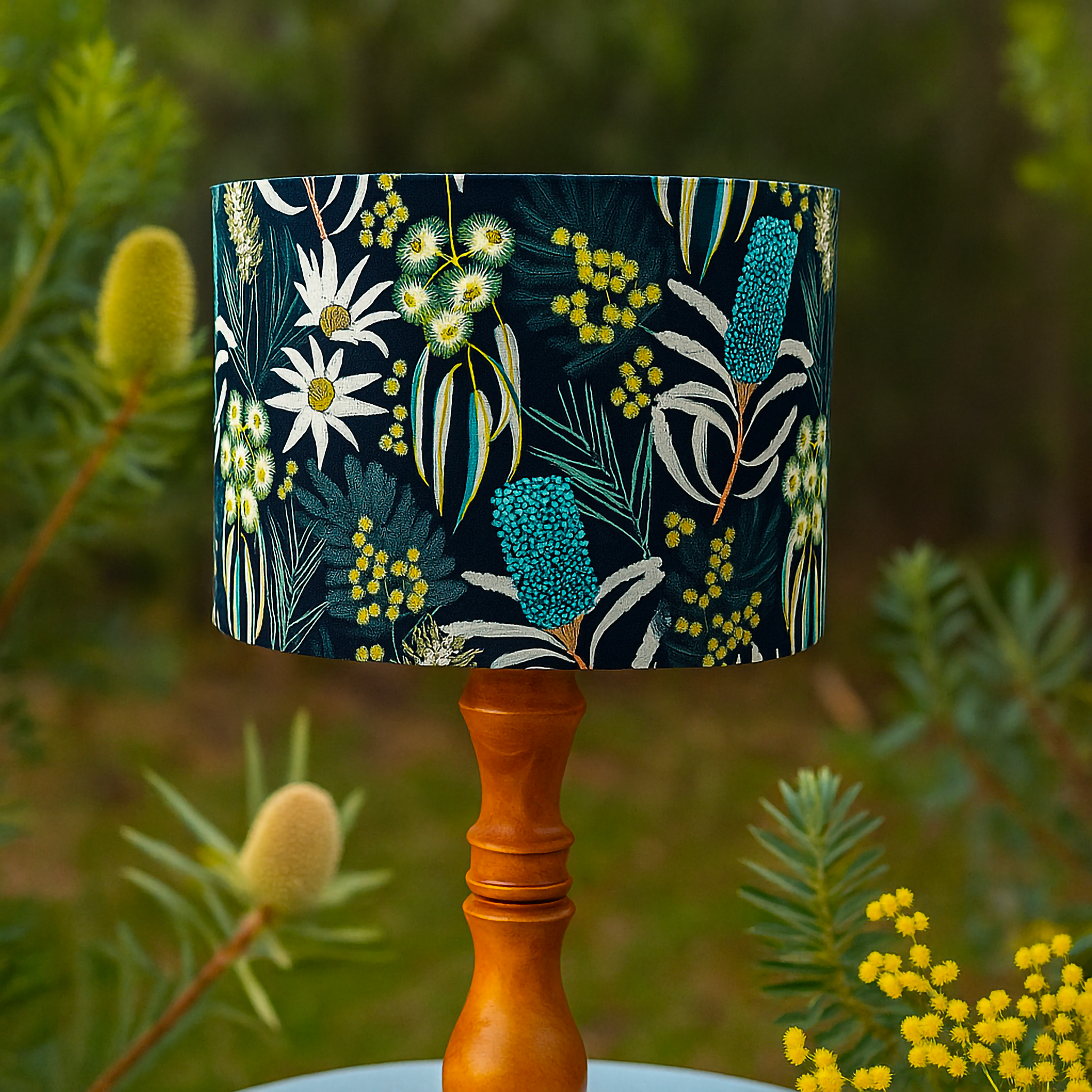 Wooden table lamp with a fabric shade featuring Australian native floral designs, including wattle, banksia, and gum blossoms in green, yellow, turquoise, and white on a deep navy background. Styled outdoors among native plants, this Luxe and Humble lampshade celebrates Australia’s botanical beauty in modern home décor.