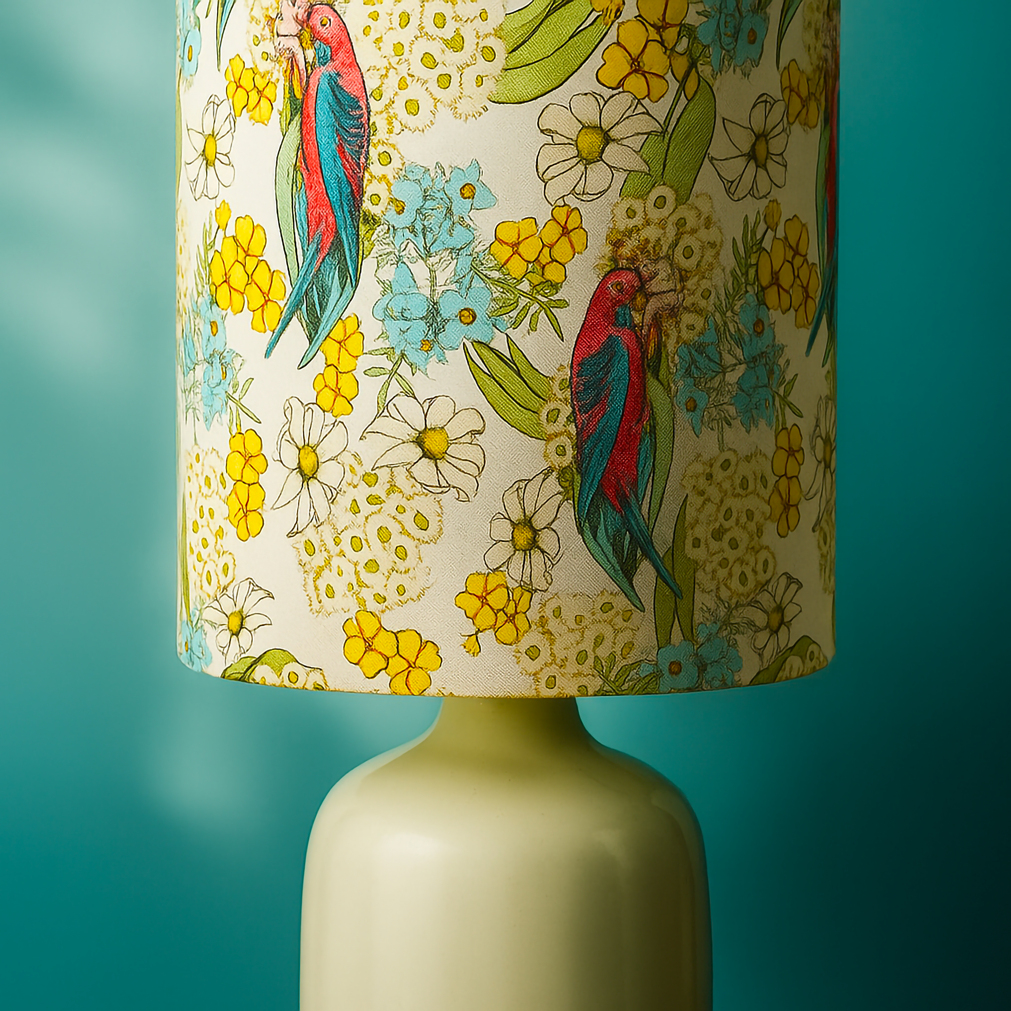 Table lamp with a fabric shade featuring colourful red and blue parrots surrounded by yellow wattle, white daisies, and soft blue blossoms. Set on a smooth cream ceramic base against a teal background, this Luxe and Humble lampshade combines native Australian birdlife with vibrant floral design for a fresh, nature-inspired home décor accent.