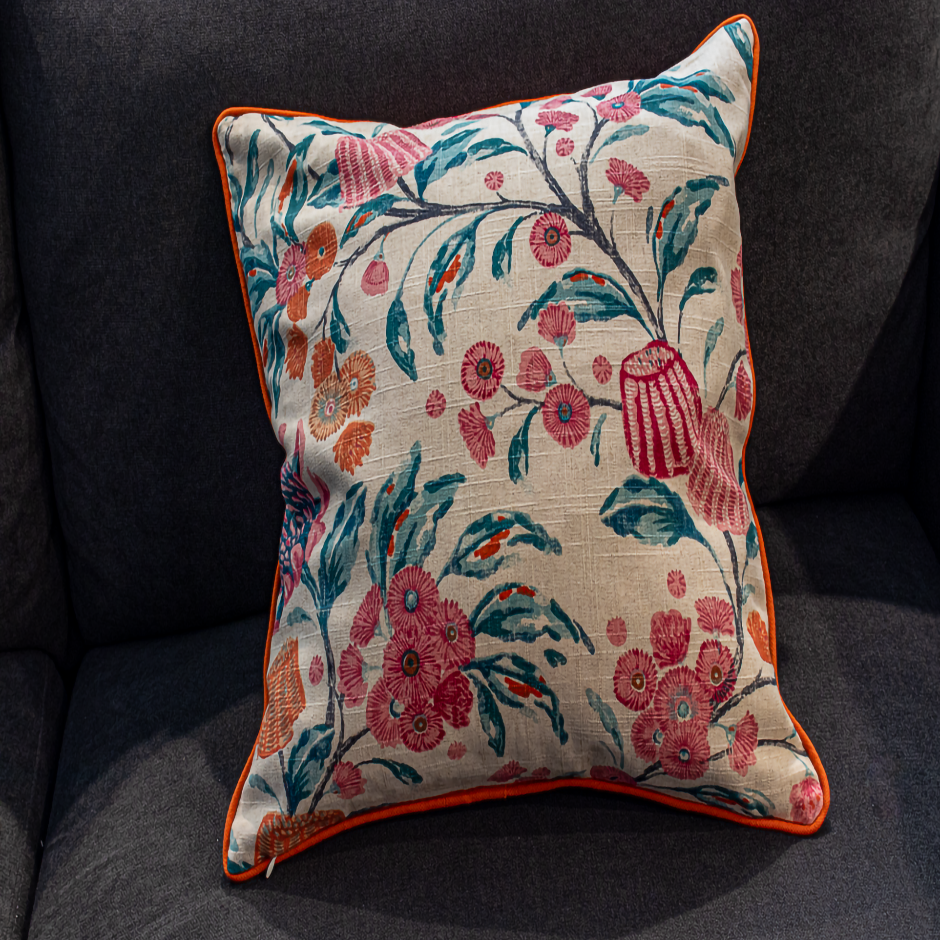 Luxe and Humble reversible cushion featuring a bold floral design in pink, coral, and teal with hand-block inspired detailing, finished with striking orange piping, styled on a dark sofa for a colourful, artisan home accent.