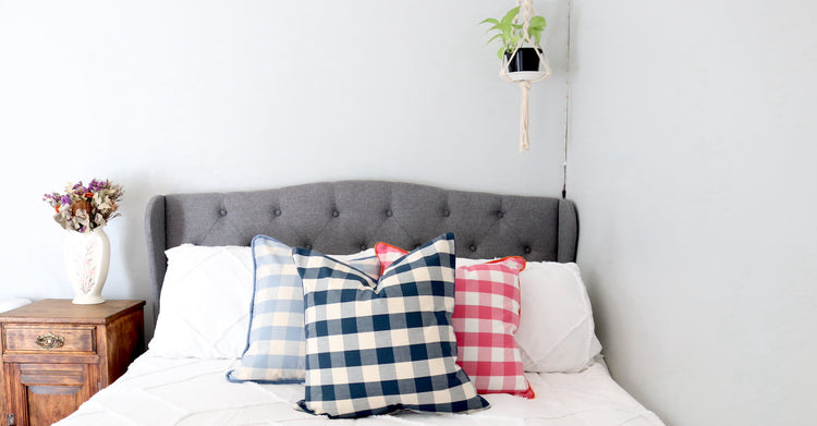 A periwinkle, pink and navy gingham cushion on a grey upholstered bed with a boho white fringed duvet cover. 