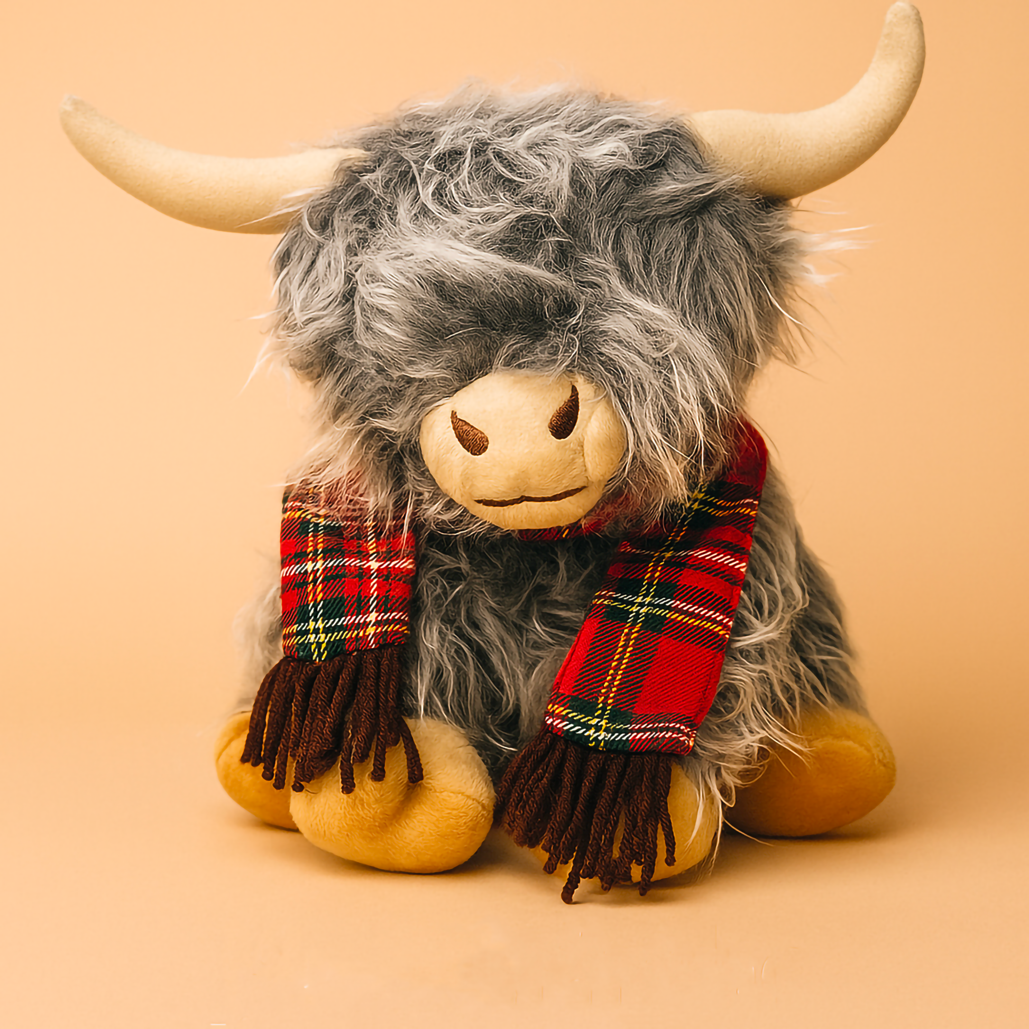 A 25cm grey Highland Cow plush toy with shaggy fur, tan horns and a red tartan scarf, photographed on a warm studio background. Cute, soft and perfect for kids’ rooms, gifting and cosy home décor. Australian boutique plush toy from Luxe & Humble.