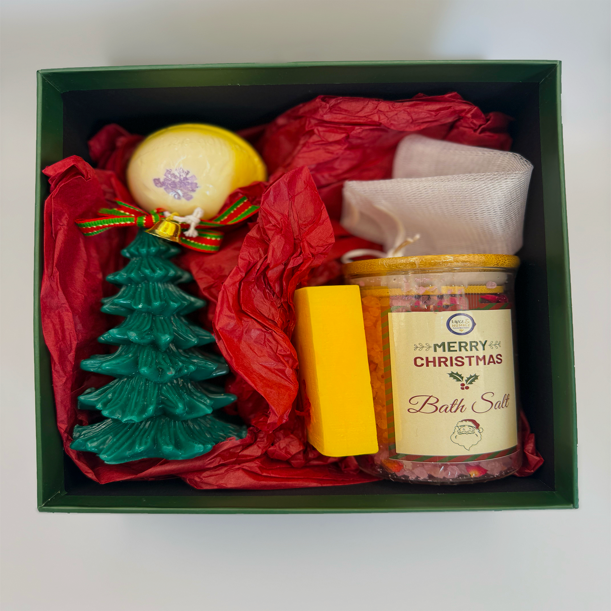 A Luxe & Humble Christmas pamper box featuring a yellow bath bomb with a purple centre, a green Christmas tree candle, a bright yellow soap bar, and a jar of colourful bath salts with a “Merry Christmas” label. Packed in red tissue inside a dark green gift box. A festive Australian self-care gift perfect for Christmas hampers, staff gifts and holiday pampering.