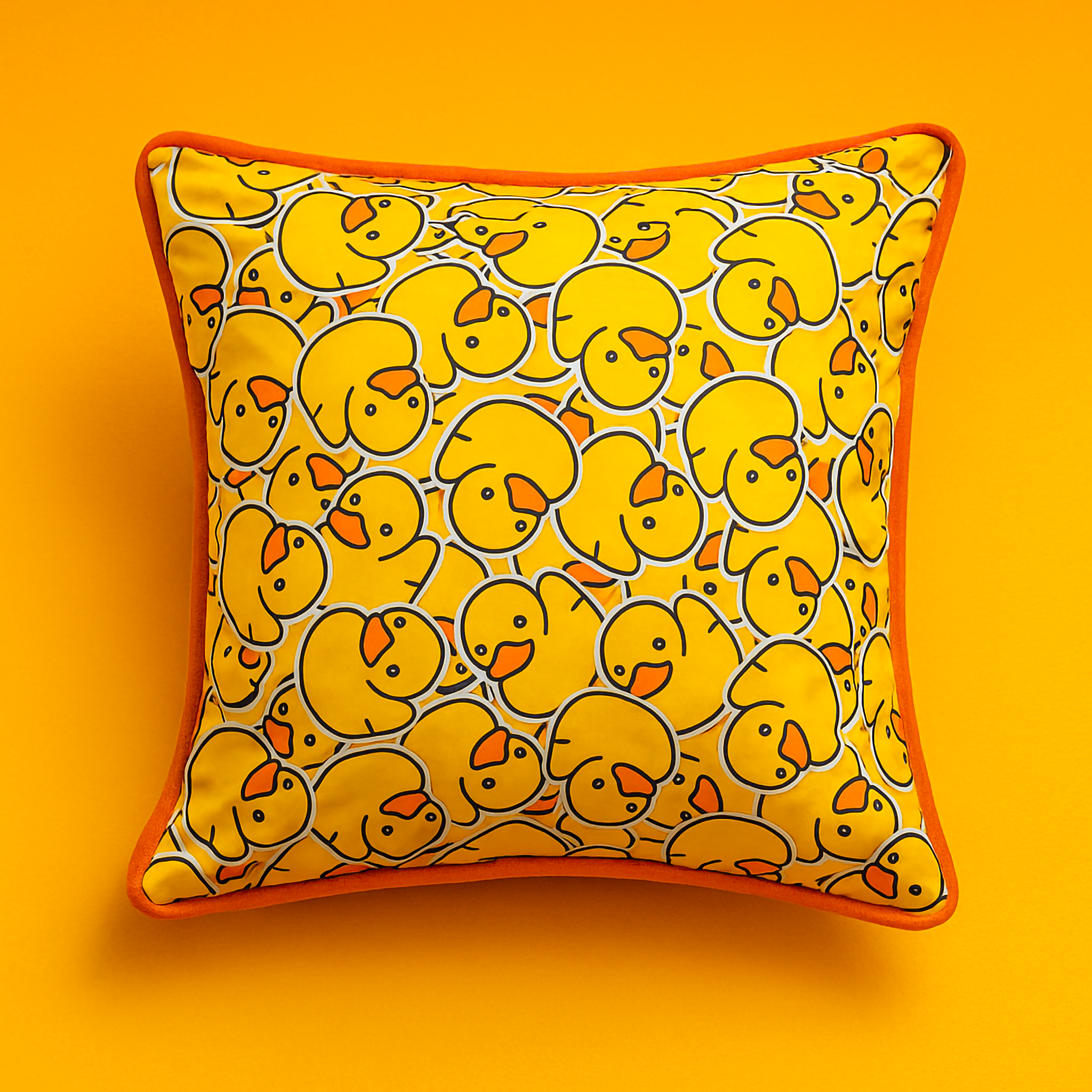 A close-up of the Duckie Print Cushion, showing the cheerful yellow duckling pattern and contrasting orange piping, displayed on a dark sofa.