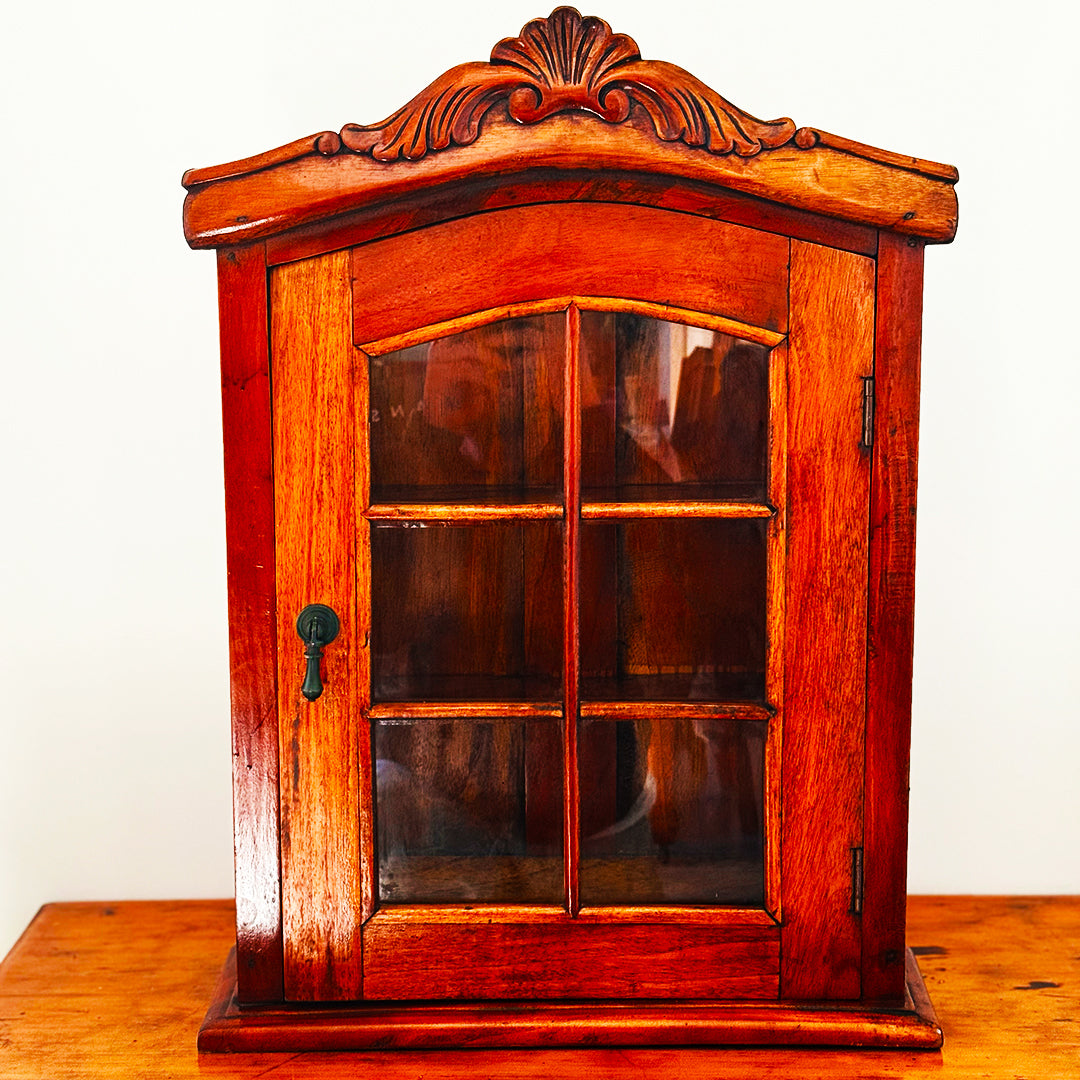 Ornate wooden wall cabinet with glass-pane door and carved arch, sitting on warm timber table