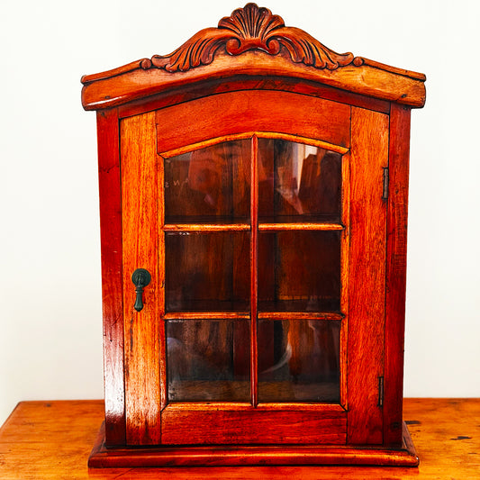 Ornate wooden wall cabinet with glass-pane door and carved arch, sitting on warm timber table