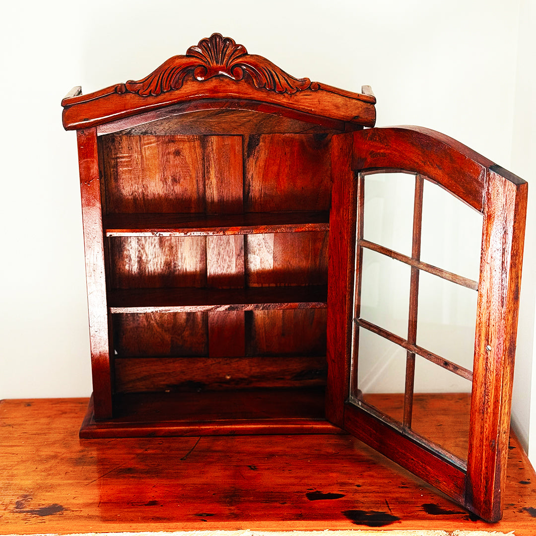 Ornate wooden wall cabinet with glass-pane door and carved arch, sitting on warm timber table