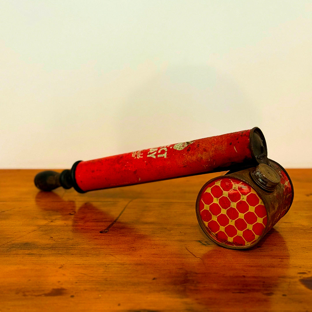 A close-up of a vintage red insect spray can, labeled "Embassy Insect Spray" with the words "Kills Household Pests" in white. The can shows signs of wear with a rustic, weathered look, and is placed on a wooden surface.

