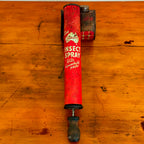 A close-up of a vintage red insect spray can, labeled "Embassy Insect Spray" with the words "Kills Household Pests" in white. The can shows signs of wear with a rustic, weathered look, and is placed on a wooden surface.