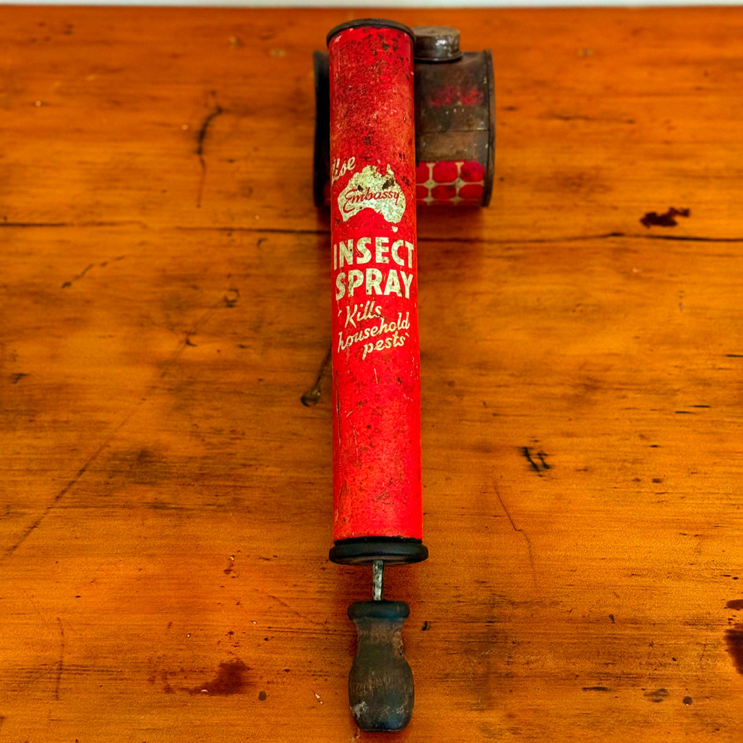 A close-up of a vintage red insect spray can, labeled "Embassy Insect Spray" with the words "Kills Household Pests" in white. The can shows signs of wear with a rustic, weathered look, and is placed on a wooden surface.