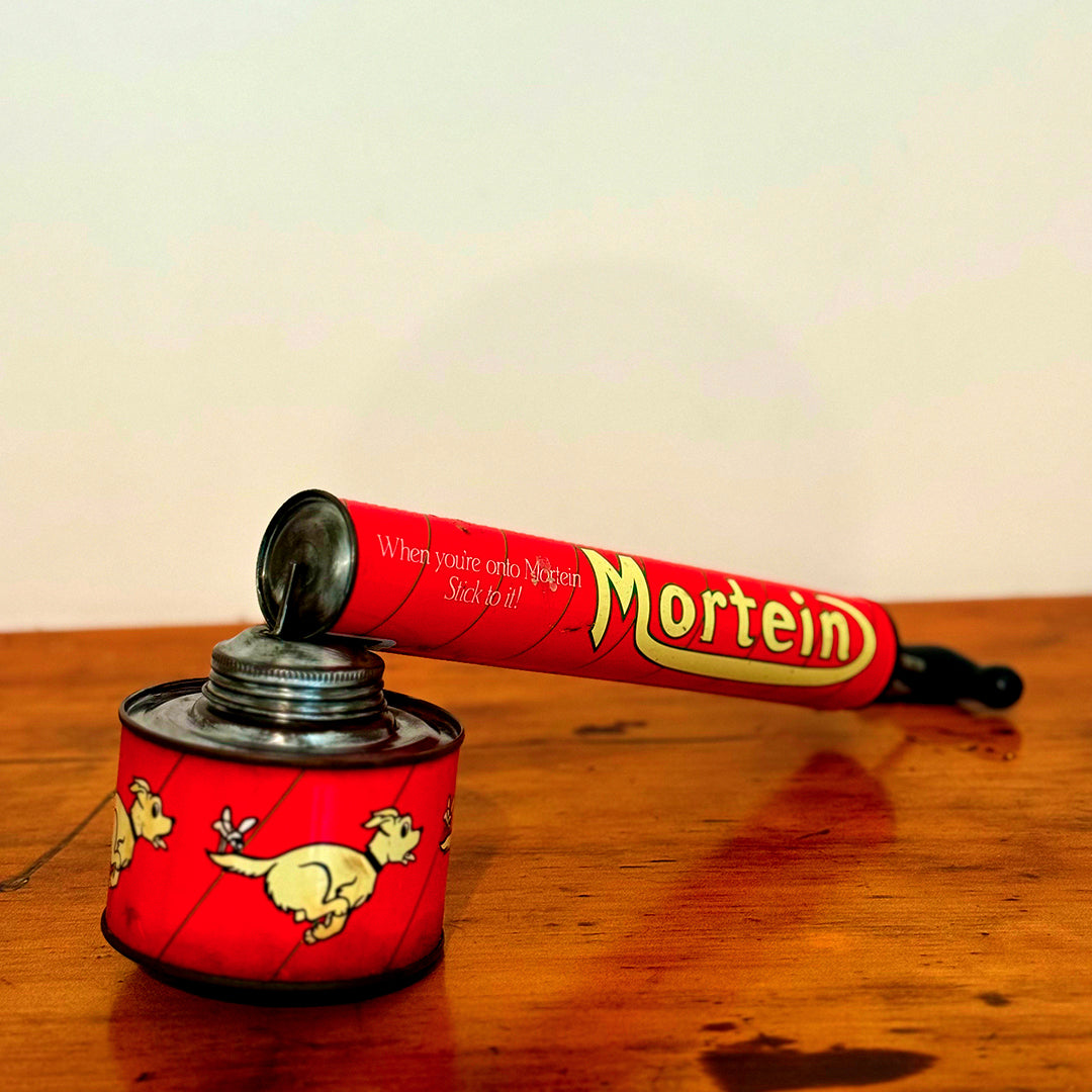 Vintage Mortein insect spray hand pump featuring a red body with the slogan "When you’re onto Mortein, Stick to it!" and classic Mortein dog logo. A retro collectible piece on a wooden surface.