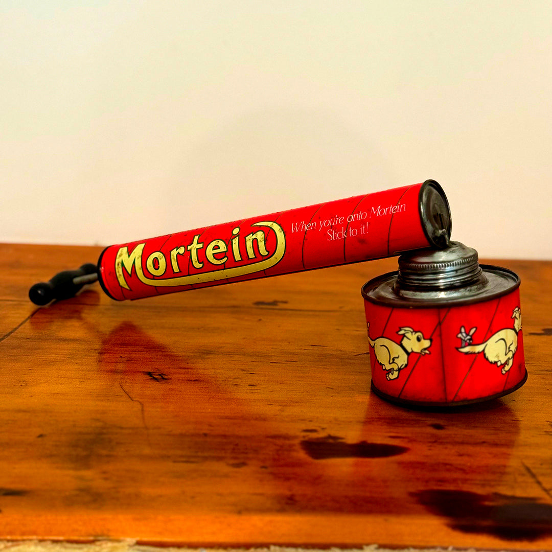 Vintage Mortein insect spray hand pump featuring a red body with the slogan "When you’re onto Mortein, Stick to it!" and classic Mortein dog logo. A retro collectible piece on a wooden surface.