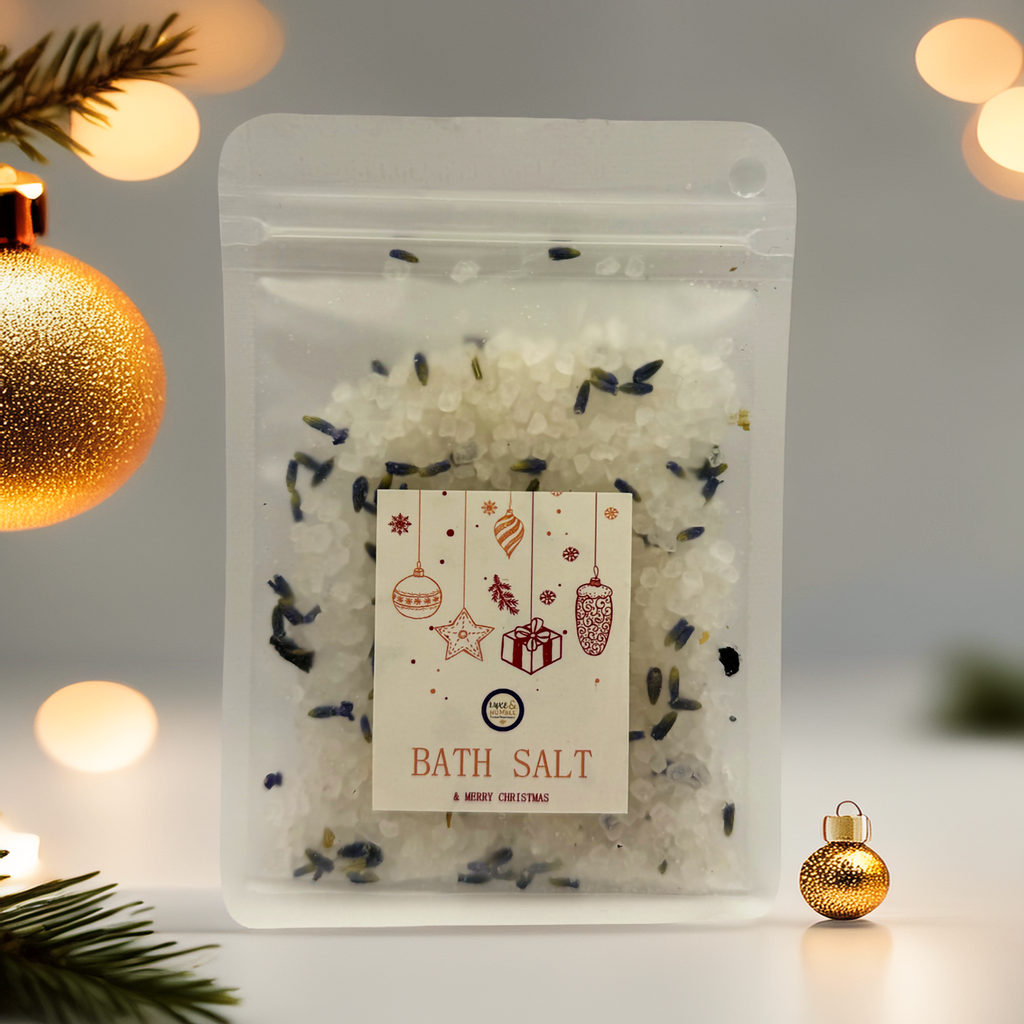 A Luxe & Humble Lavender Snow Christmas Bath Salt pouch filled with white mineral salts and dried lavender buds, displayed in a clear stand-up bag with a festive Christmas label featuring hanging ornaments and gift illustrations. Styled with soft golden baubles, warm bokeh lighting and pine sprigs — perfect for Australian Christmas gifting, stocking fillers and self-care.