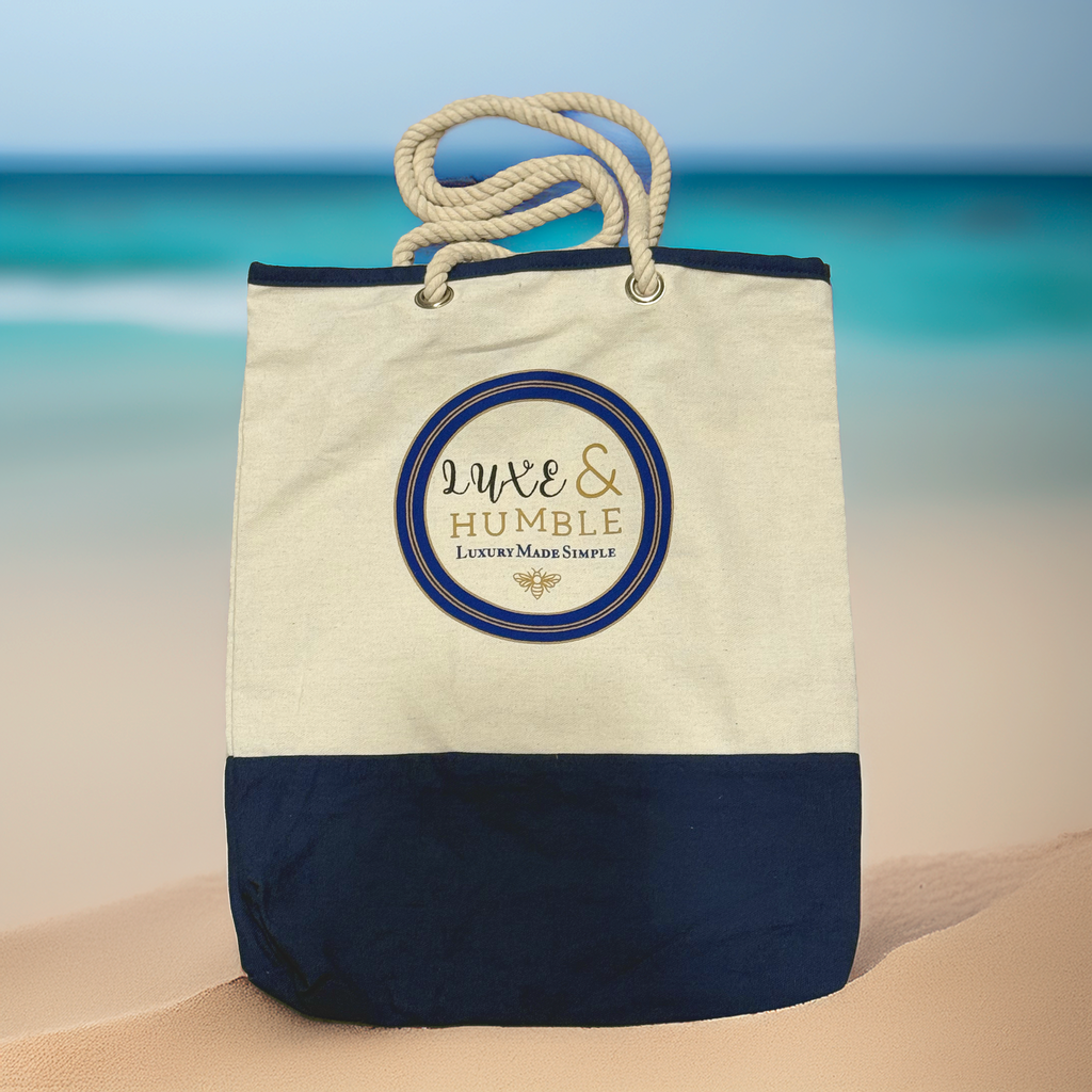 A Luxe & Humble navy and natural canvas tote bag with rope handles displayed upright on soft sand with a blurred turquoise ocean backdrop. Premium Australian coastal lifestyle image showcasing the bag’s texture and signature logo.
