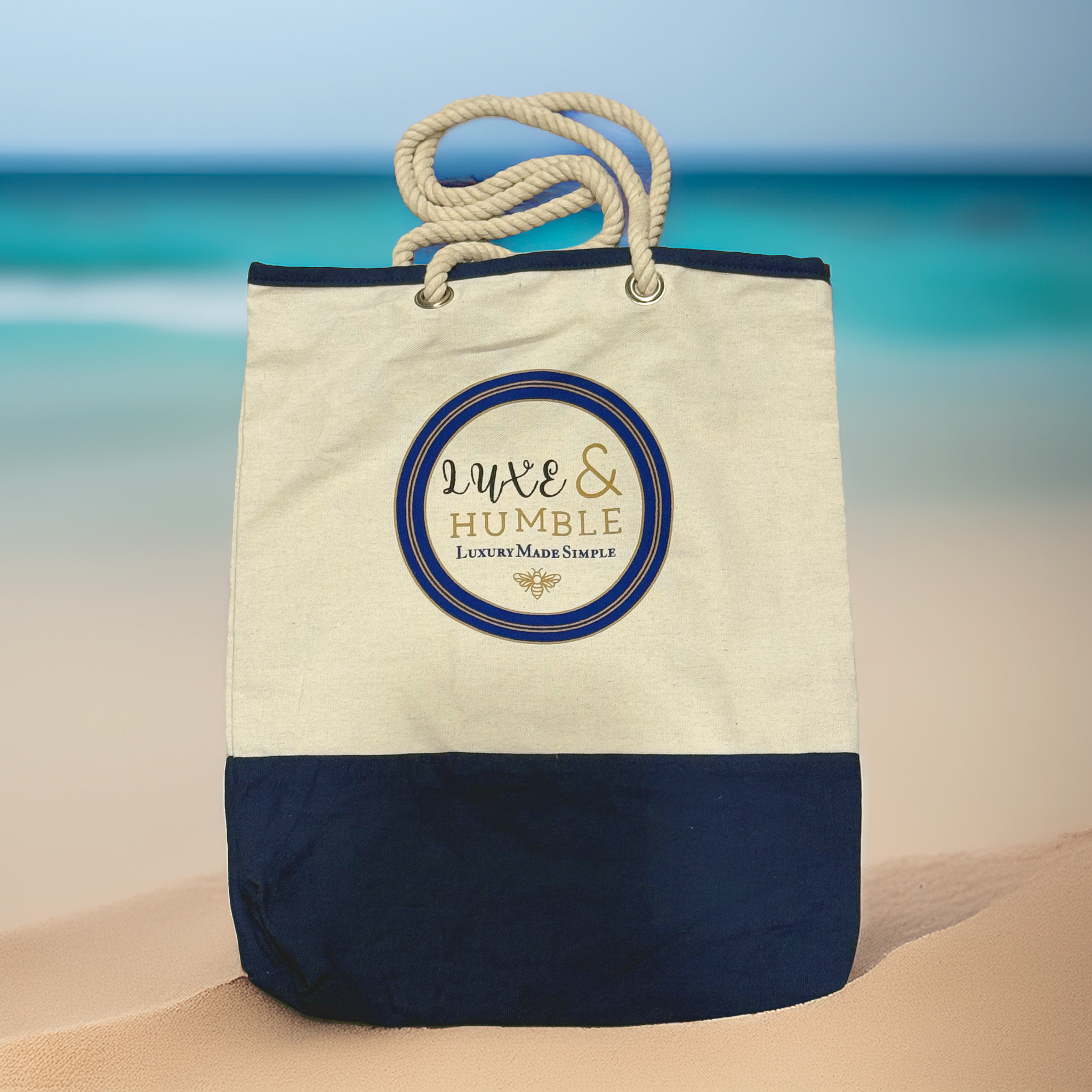A Luxe & Humble navy and natural canvas tote bag with rope handles displayed upright on soft sand with a blurred turquoise ocean backdrop. Premium Australian coastal lifestyle image showcasing the bag’s texture and signature logo.