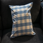 A close-up of the Luxe & Humble Gingham Blue and Beige Pillow featuring a soft cotton texture and a classic checkered gingham pattern on a dark sofa.
