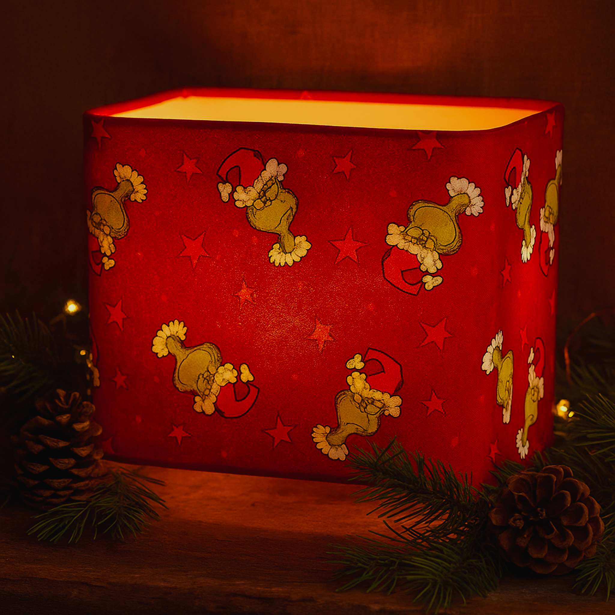 Handmade Luxe & Humble rectangular 20 x 40 cm lampshade featuring red cotton fabric with playful Grinch Santa print and star motifs, glowing warmly on a rustic wooden shelf.