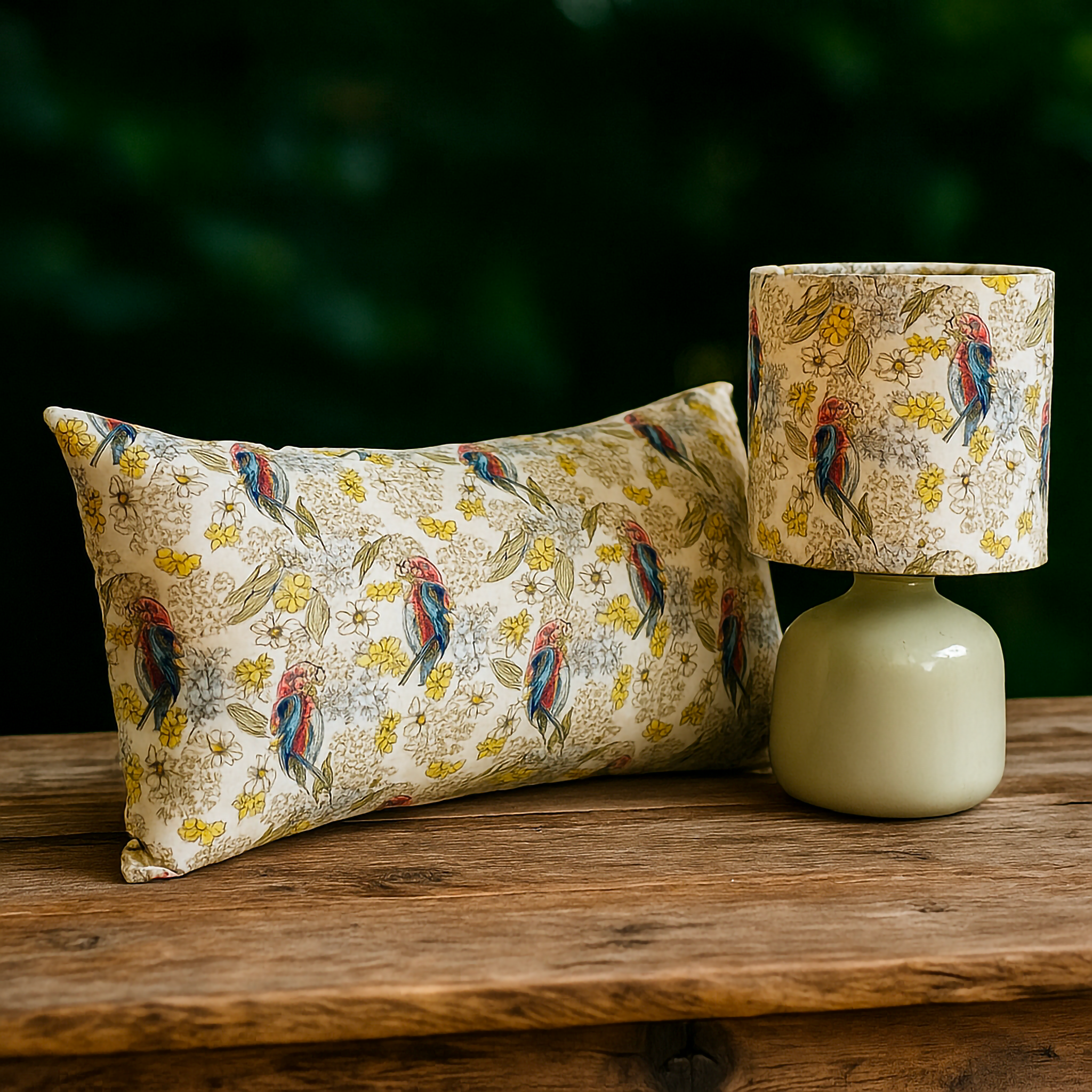 Rectangular cushion and matching lampshade with fabric design featuring red and blue Australian parrots surrounded by yellow wattle and delicate white flowers. Styled outdoors on a rustic wooden surface with a dark green natural backdrop, this Luxe and Humble décor set blends native birdlife with floral elegance for a fresh and vibrant home accent.