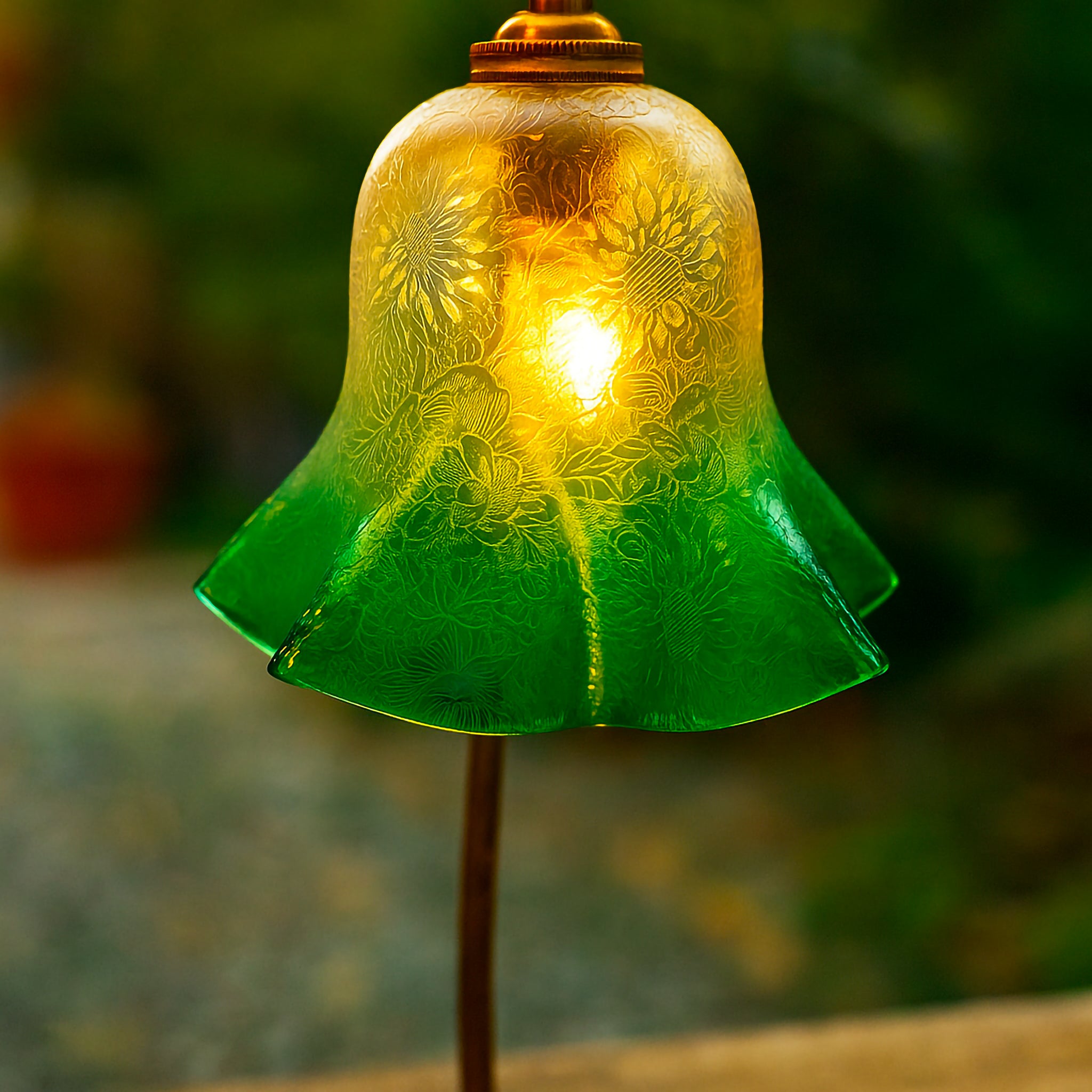 Close-up of a bell-shaped glass lampshade with intricate etched floral patterns, blending clear and green tones. Mounted on a brass fitting and styled outdoors with soft greenery in the background, this Luxe and Humble piece highlights fine craftsmanship and timeless decorative lighting design.