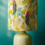Table lamp with a fabric shade featuring colourful red and blue parrots surrounded by yellow wattle, white daisies, and soft blue blossoms. Set on a smooth cream ceramic base against a teal background, this Luxe and Humble lampshade combines native Australian birdlife with vibrant floral design for a fresh, nature-inspired home décor accent.