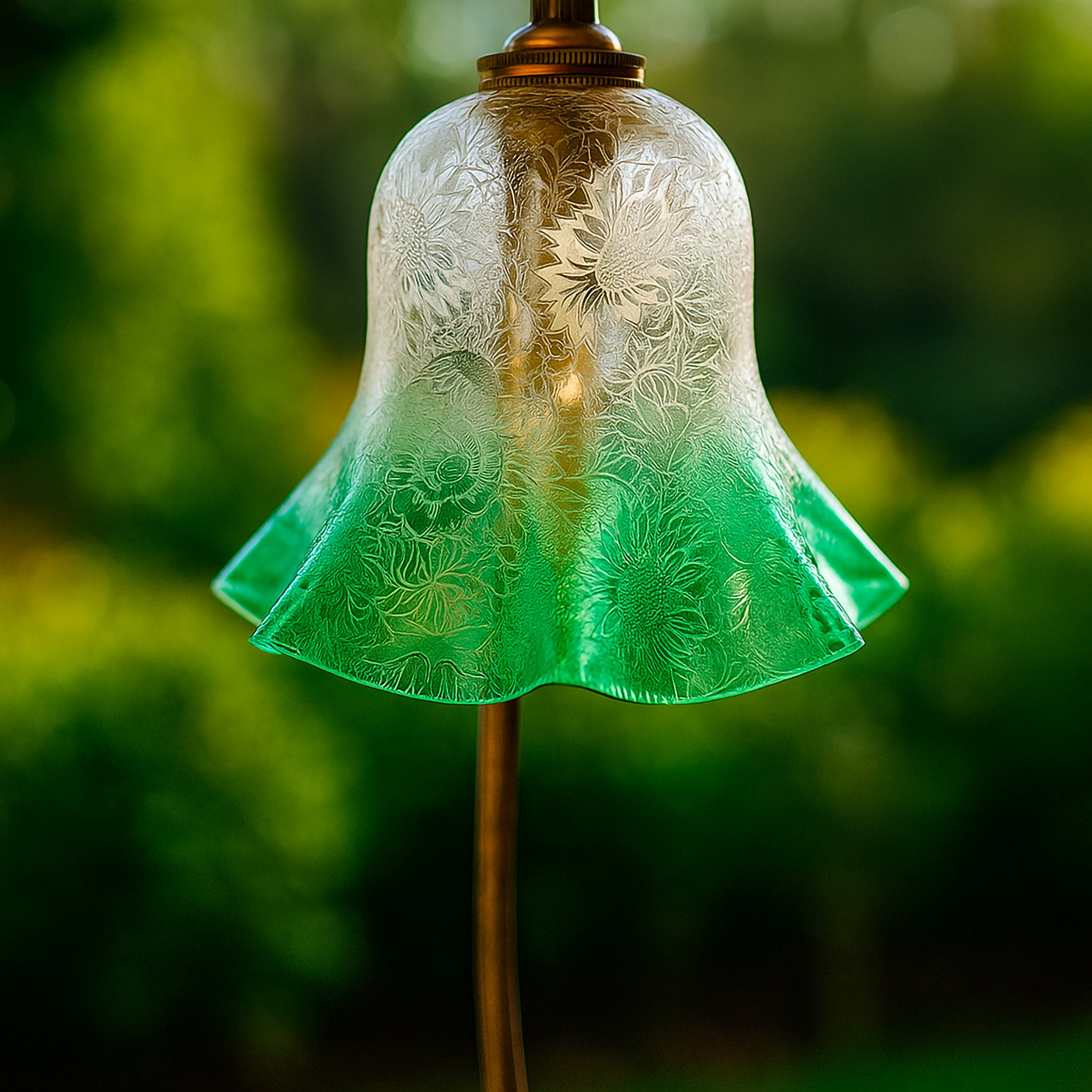 Close-up of an elegant glass lampshade in golden yellow and emerald green tones with delicate etched floral patterns, glowing warmly with light. Styled outdoors with a soft natural background, this Luxe and Humble piece highlights craftsmanship and artistry in decorative lighting.