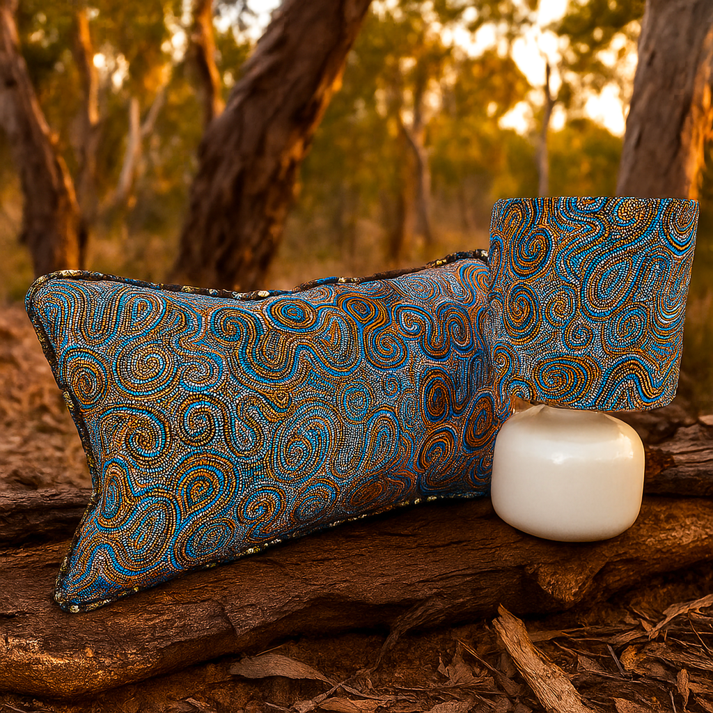 Rectangular cushion and matching lampshade featuring intricate Indigenous-inspired dot and swirl artwork in blue, orange, and earthy tones. Styled outdoors on a tree log with a native Australian bushland backdrop, this Luxe and Humble décor set blends cultural artistry with natural elegance.