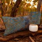Rectangular cushion and matching lampshade featuring intricate Indigenous-inspired dot and swirl artwork in blue, orange, and earthy tones. Styled outdoors on a tree log with a native Australian bushland backdrop, this Luxe and Humble décor set blends cultural artistry with natural elegance.