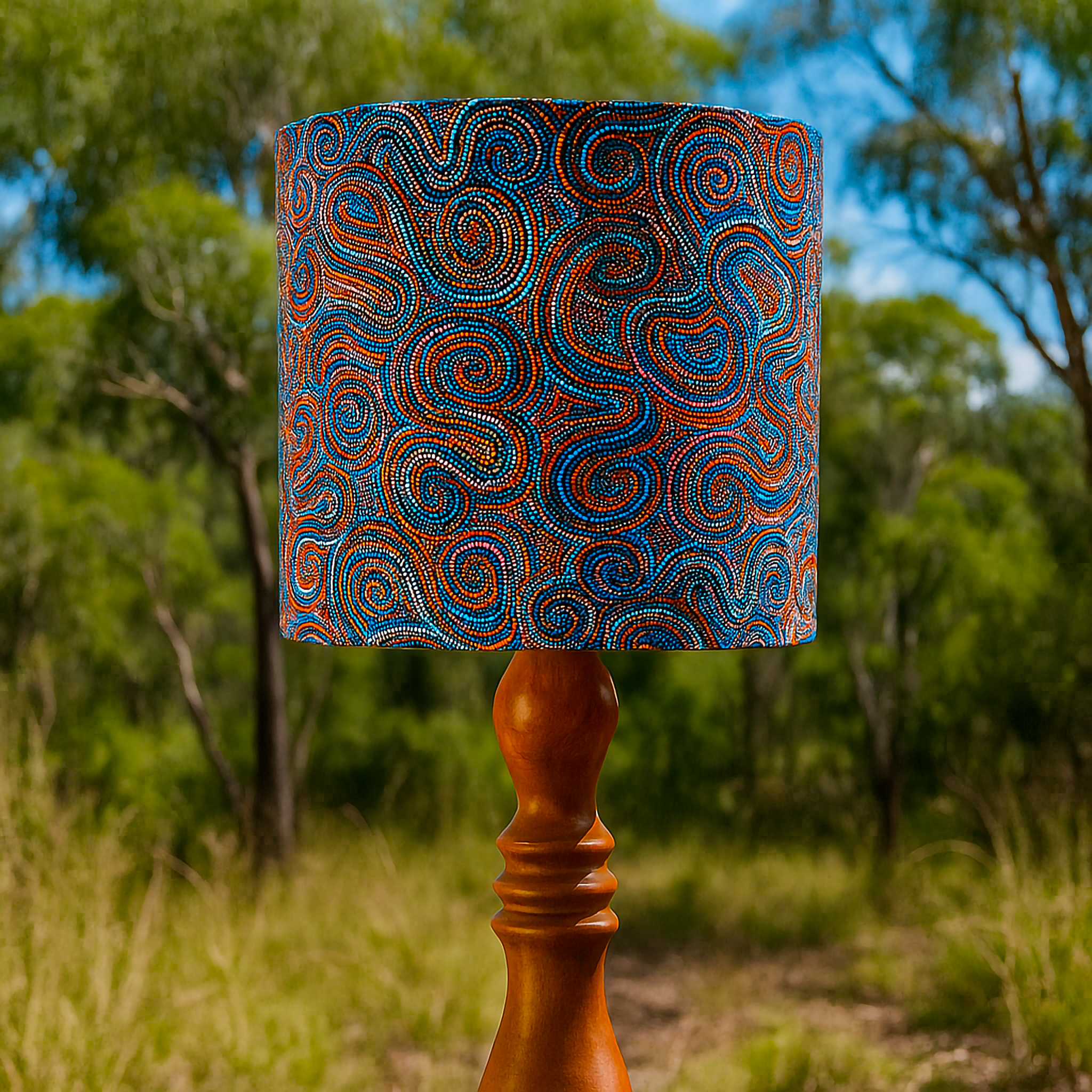 Wooden table lamp with a fabric shade featuring ‘Whirlwind Country’ dot artwork by acclaimed Warlpiri artist Pauline Napangardi Gallagher. The swirling blue, orange, and red dotted patterns capture the Mina Mina Jukurrpa (Dreaming), styled outdoors with Australian bushland in the background. A Luxe and Humble décor piece blending authentic Aboriginal storytelling with contemporary home design.