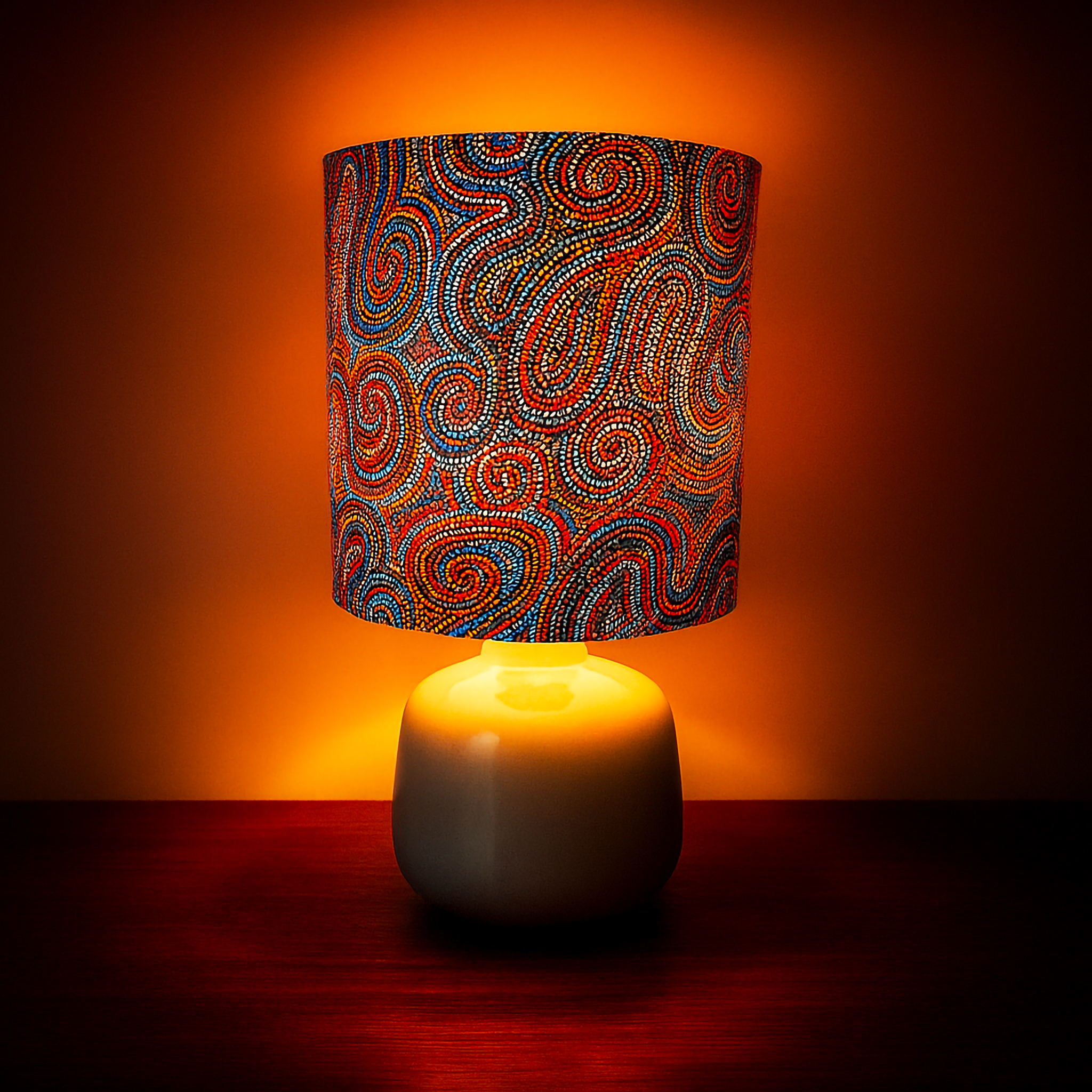 Table lamp featuring ‘Whirlwind Country’ dot artwork by Pauline Napangardi Gallagher. The vibrant fabric shade showcases intricate swirls in blue, red, and orange tones, paired with a white ceramic base. Styled with warm lighting, this Luxe and Humble décor piece blends authentic Indigenous art with contemporary home design.