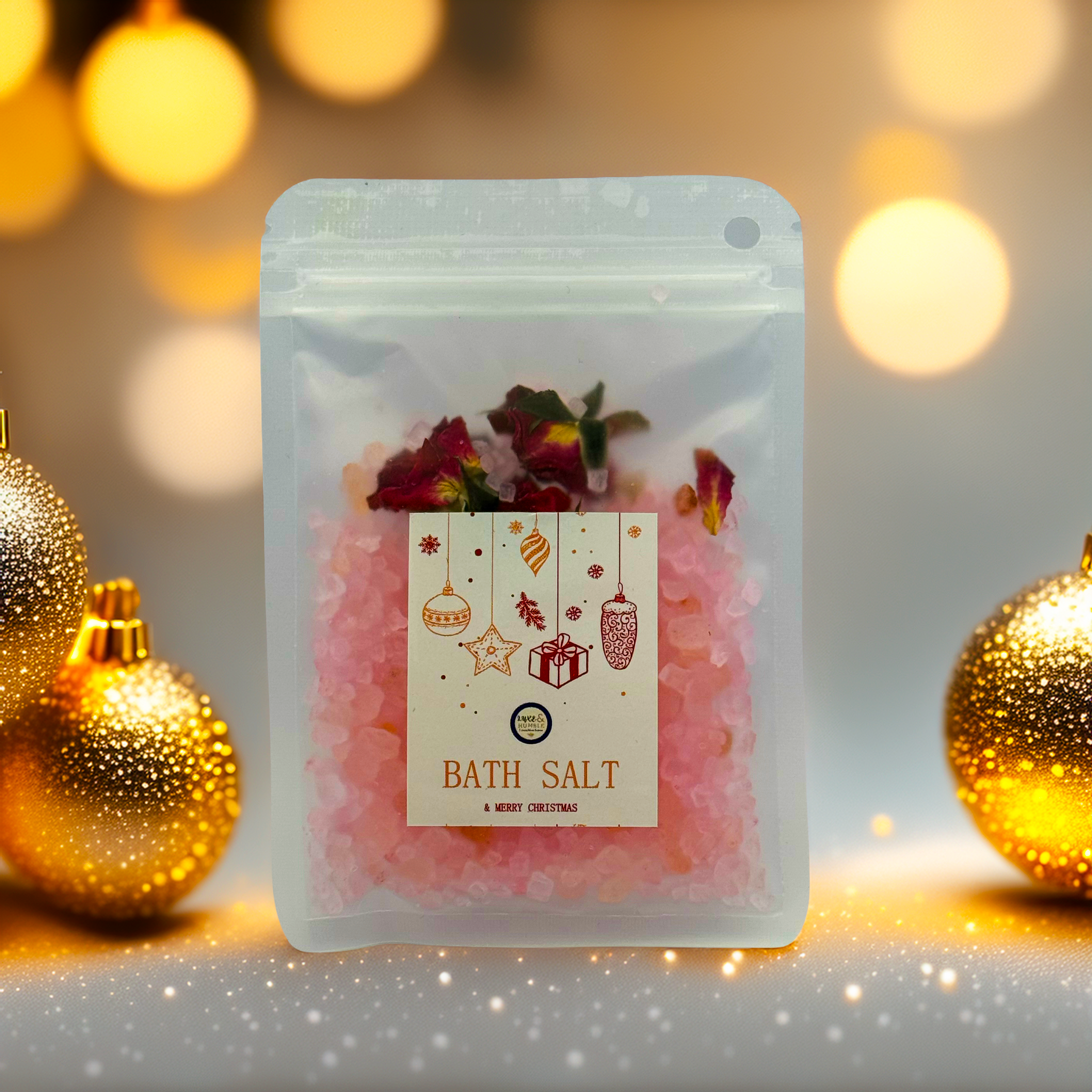 A Luxe & Humble Rose Blush Christmas Bath Salt displayed in a clear resealable pouch filled with pink mineral salts and dried rose petals. The festive front label features hanging Christmas ornaments, stars and a gift illustration. Styled with glowing golden baubles, warm bokeh lights and soft sparkles, this product image captures a luxurious and cosy holiday feel, perfect for Australian Christmas gifting, stocking fillers and self-care lovers.