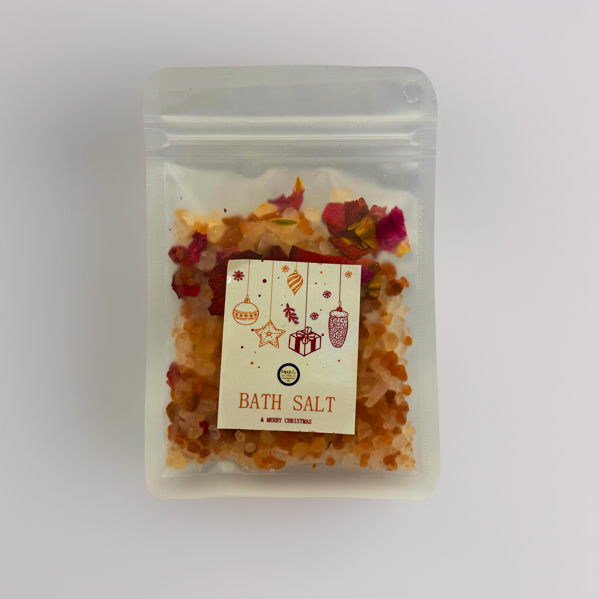 A Luxe & Humble Sunset Rose Christmas Bath Salt displayed in a clear stand-up pouch filled with orange crystal salts and rose petals. Styled with glowing golden Christmas baubles, pine sprigs and soft festive sparkles. A handcrafted Australian bath salt perfect for Christmas gifting, Secret Santa and holiday pampering.