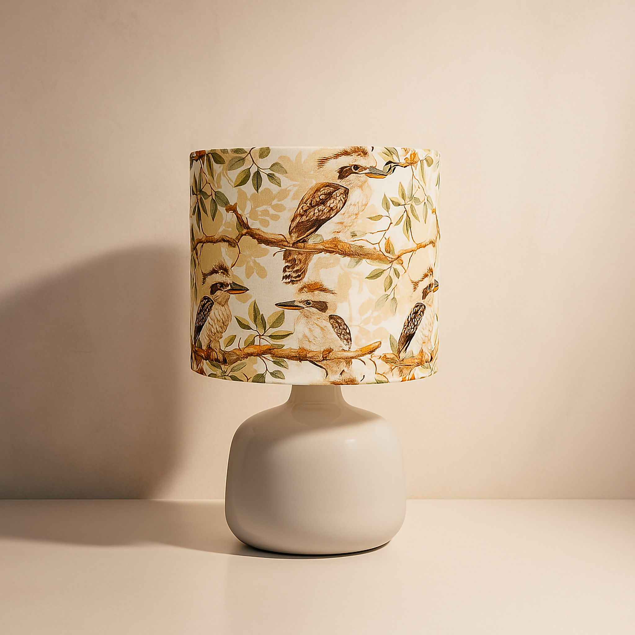 A handcrafted table lamp by Luxe & Humble featuring a 15x15cm drum lampshade printed with Australian kookaburras perched on eucalyptus branches. The lamp sits on a smooth 12.5x12.5cm gloss ceramic base, creating a warm, nature-inspired lighting piece that celebrates Australian wildlife and bushland charm. Perfect for modern country interiors, bedrooms, or living spaces.