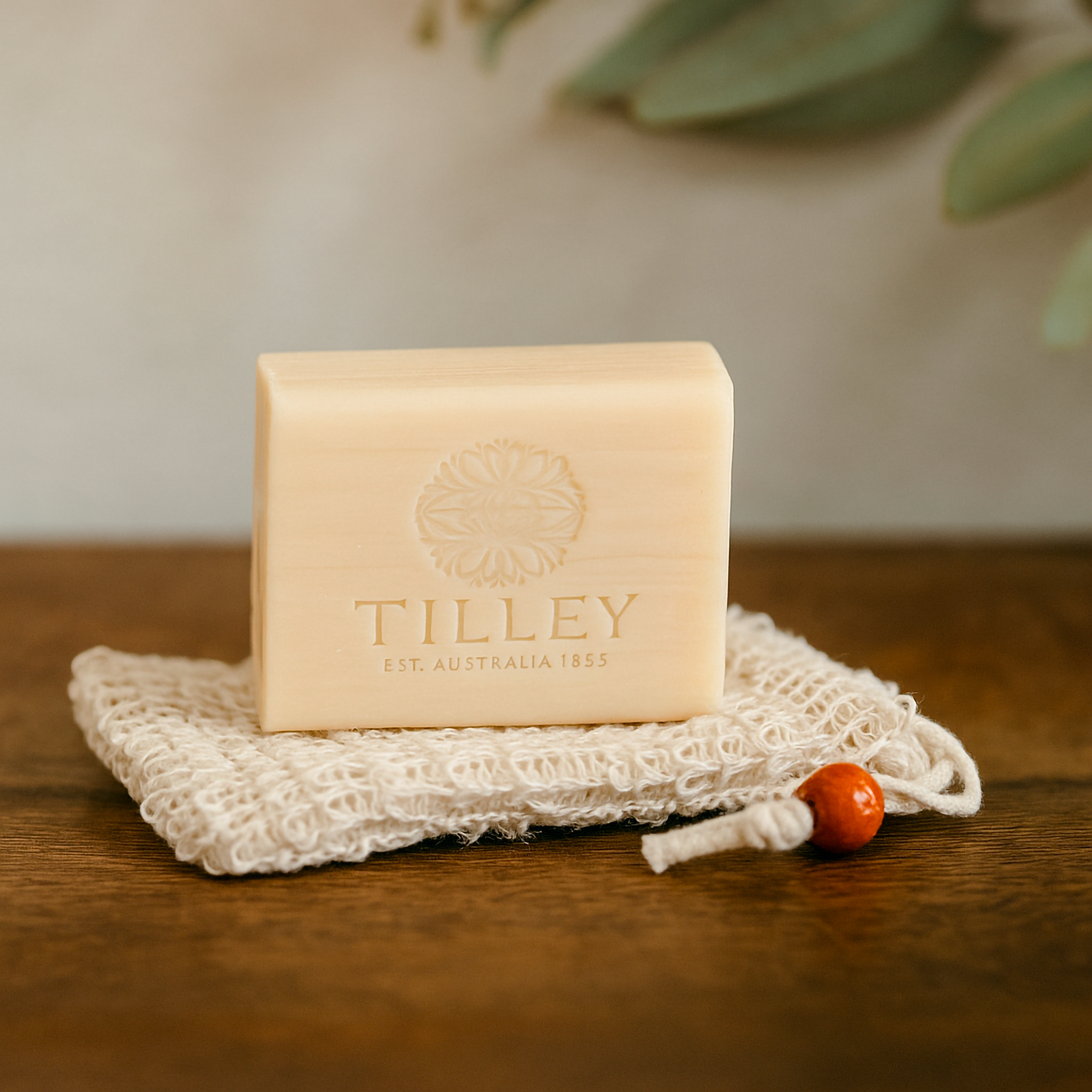 A Luxe & Humble Purity Pouch soap saver made from natural sisal fibres with wooden bead drawstring, displayed with a Tilley soap bar on a wooden surface beneath soft greenery — evoking calm, Australian-made luxury.